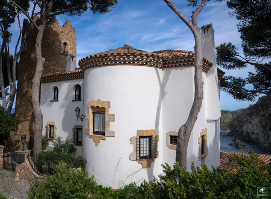Color photo of a tile roofed white stucco house with a rounded turret on the near side and a square stone tower on the far side. In the background below can be seen a peek of the waters of a cove or cala. 
