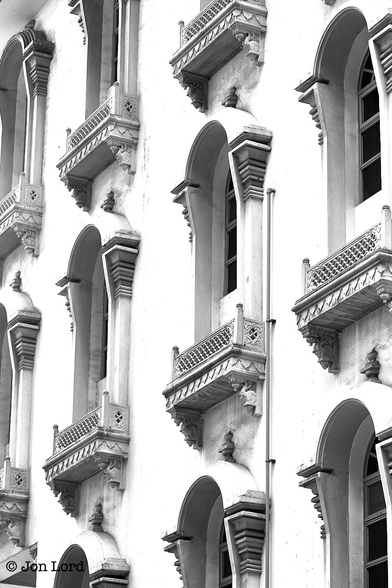 This is a black and white street / architectural photo in portrait format of the facade of a period city building with numerous windows all surrounded by an masonry arch that extends in front of the opening. Waterloo Street, Singapore (2013).

The camera is tilted upwards at the off-white masonry facade of a period urban building. The view is oblique with the camera angled at 45º to the right of the front and viewing along the length of the building.  There are 3 floors in view and with the camera's perspective: the windows tilt downwards from the right to the left. Only the centre floor windows are completely shown, the upper and lower are cropped by the top and bottom edges of the photo. All the windows have an masonry arch surrounding each opening and extending outwards about 30cm. There is a tall pillar on either side of the window with the arch circling the top and with a small spike on the crest of the arch. At the base is a delicately decorated window box (devoid of flowers) with small ornately carved elephants heads supporting the box at each end. 

The building is the small block immediately behind and attached to the Sri Krishnan Temple. The carved elephants heads perhaps suggesting the connection of the building to the Hindu faith.