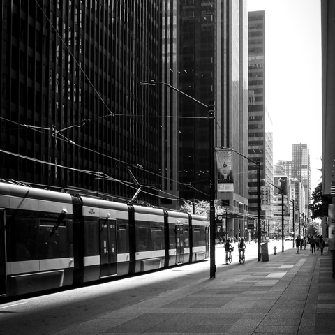 A streetcar is seen passing by tall glass windowed buildings on a busy city street. People can be seen in the distance walking along the wide sidewalk and a couple of bicyclists are on the road. This is a black and white image.