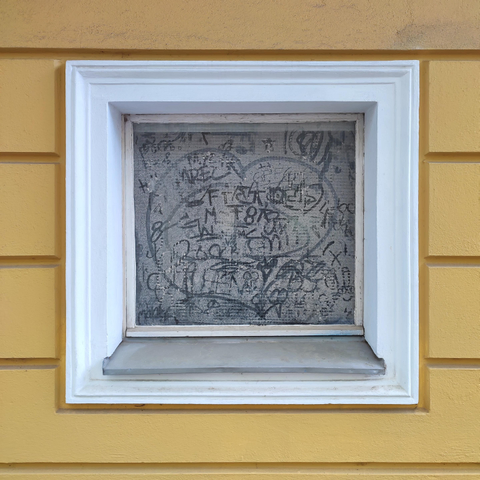Quadratisches Farbfoto. In einer gelb verputzten Wand ist ein kleines Fenster mit weißem Fensterrahmen eingelassen. Die Scheibe ist sehr staubig, verschiedene Menschen haben mit dem Finger Zeichnungen und Buchstaben in den Staub gezogen. In der Mitte ist ein großes Herz.