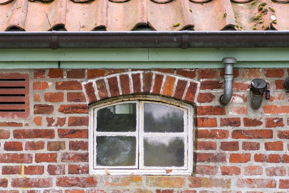 Fenster mit weißem Rahmen und vier Scheiben in einer roten Backsteinwand unter einem Dach mit roten Ziegeln