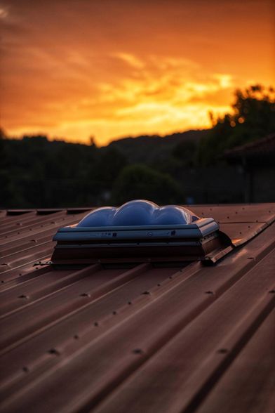 Das Bild zeigt eine stimmungsvolle Abendaufnahme eines Metalldaches während eines farbenprächtigen Sonnenuntergangs. Im Zentrum des Motivs befindet sich eine moderne, kuppelförmige Lichtkuppel aus bläulich schimmerndem Kunststoff, die auf einem dunklen, trapezförmigen Blechdach montiert ist. Die Dachfläche führt den Blick perspektivisch von unten rechts nach oben links in die Tiefe. Über der Silhouette bewaldeter Hügel am Horizont erstreckt sich ein dramatischer Himmel, der in intensiven Orange-, Gold- und dunklen Rottönen leuchtet. Das warme, indirekte Licht der untergehenden Sonne spiegelt sich sanft auf den Erhöhungen des Profildaches wider und erzeugt lange, weiche Schatten, während die Details am unteren Bildrand in einer ruhigen Dunkelheit verschwimmen.