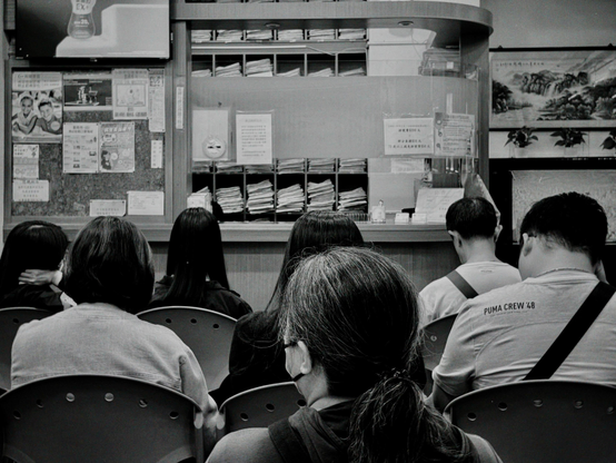 People sitting in chairs facing a counter or window with notices, in an indoor setting.