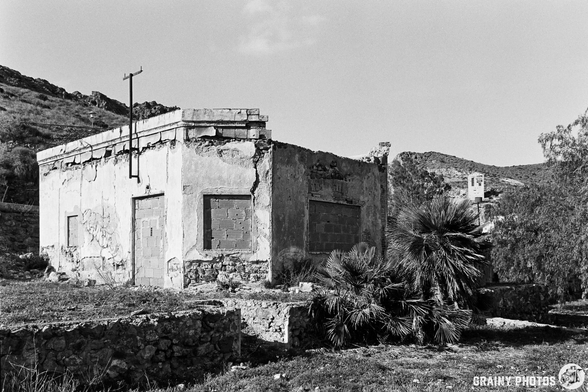 Abandoned structure on a hillside, showing signs of decay and overgrowth. The scenery includes rocky terrain and sparse vegetation, with remnants of past architecture in a desolate landscape. Black and white photography enhances the atmosphere of neglect.