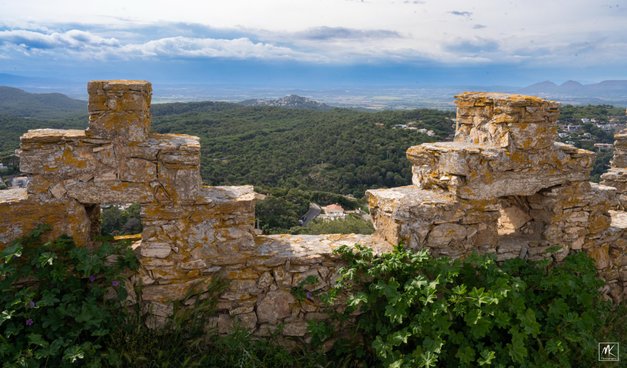 Color photo with castle battlements in the shape of stepped pyramid-shaped stonework in the foreground and beyond them the hills and plains of Catalonia’s Baix Empordà interior landscape.