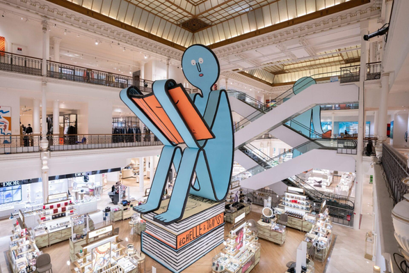 an enormous blue illustrated figure reading an orange book in the middle of a mall