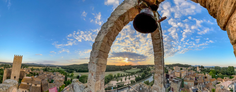 Panoramic photo taken near sunset from atop a bell tower in a medieval Catalonian town.  The bell is suspended beneath an arch formed by three spindly piers. 