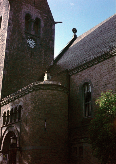 A grainy photograph of a the asymmetric tower and entrance to a stone church from the early 1900s. The photo was taken against the sun, so most of the church is in the shade, with a bright blue sky in the background. The photo is a little underexposed, and the expired film has made the colours in the shadows a little funky.