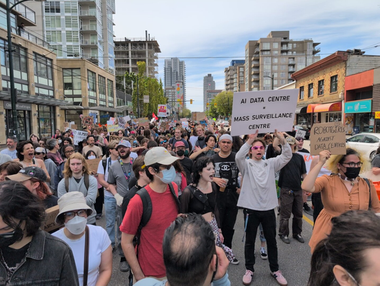 A crowd of people walking down Broadway Street in Vancouver, holding a variety of signs in protest against AI and AI data centres. In the foreground two signs are legible: "AI Data Centre = Mass Surveillance Centre," and "Robotgirls Against Data Centres."