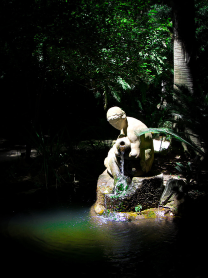 A classic stone nymph statue pouring water into a pond. Sunlight illuminates the sculpture while dense, tropical garden trees fill the background with dark shadows.