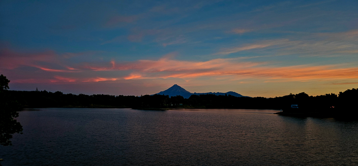 View across Lake Rotomanu in New Plymouth, Mount Taranaki peeking above the trees in the distance, and the sunset lighting up the thin strips of cloud in the blue sky with a pink glow.