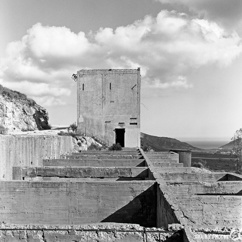 A black-and-white image of an abandoned building situated on a rocky hillside, surrounded by concrete foundations. In the background, a vast landscape extends to the horizon, where the sky meets the sea under a cloudy sky.