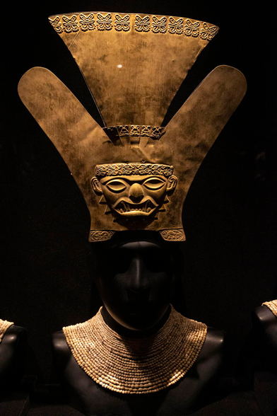 A museum exhibit shows a decorative headpiece and necklace of an Inca person of some importance. The items are mounted on a black mannequin torso in a black-backed display cabinet with a main light source falling from above and in front so that the only hint of a face on the mannequin comes from a slight highlight on the nose and forehead. The necklace comprises twenty one concentric rings of small, pale, rounded stones threaded together to lie around the neck and spread out onto the shoulders. The headpiece is a largely flat sheet of gold standing upright on the head. The shape is a little like a hand with thumb and little finger spread apart and the central fingers joined together but flaring outwards at the top. Engraved into the top ridge of the piece are butterfly shapes. At the bottom, just above where the headpiece sits on the head of the mannequin, there is an embossed, stylised face of a fierce Inca warrior. The face is wide with large, oval eyes, a thick nose, and wide, thick lips in a grimace.