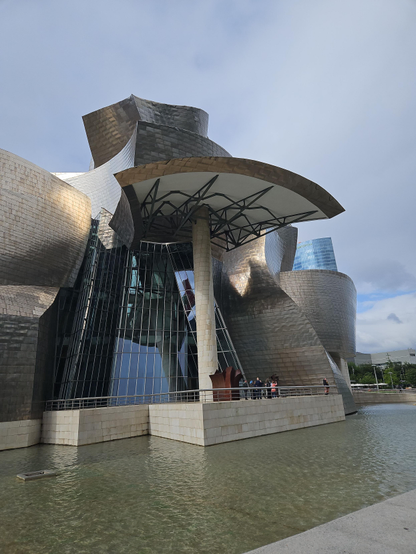 Ein großes Gebäude, das komplett zusammengepuzzelt aussieht. Säulen, Glas und geschwungene Bauteile bilden ein futuristisch aussehendes Konstrukt ohne gerade Linien und rechte Winkel. Die sichtbare Oberfläche der Wände glänzt metallisch im Licht. Zwischen dem Gebäude und dem Fotografen ist eine künstliche flache Wasserfläche in einem grauen Becken.
