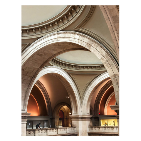 A view of some of the massive limestone arches inside the Great Hall of the Metropolitan Museum of Art in NYC, as seen from the upper floor looking across to the balcony on the far side of the main space. A large arch in the foreground rises up to meet the ornamented rim of one of the saucer-shaped domes in the hall’s vaulted ceiling. Through the arch we see two more arches underneath the rim of another dome, and through those we see even more archways framing some glass exhibit cases or leading into other spaces. A person pushing a stroller walks alongside the balcony railing, dwarfed by the architecture. The lighting gives the spaces under the far archways a soft, warm glow.