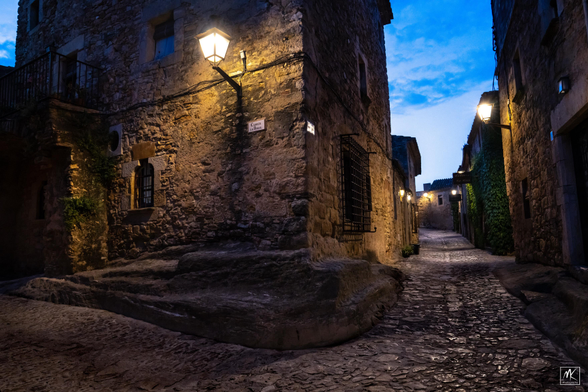 Color photo of an old stone building at a corner on intersecting cobblestone streets.  The photo was taken just after sunset during the “blue hour” with dark, partly cloudy blue skies above and the streets and buildings are lit by wall-mounted lamps. 