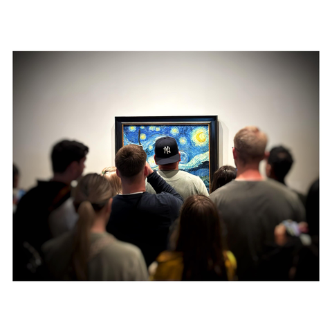 Photo of many museumgoers crowding around Vincent van Gogh’s “The Starry Night,” which hangs in a black frame on a white wall. The crowd partly obstructs our view of the iconic painting, particularly a man standing squarely in front of it wearing a backwards dark blue baseball cap with the New York Yankees logo — his head perfectly in the center of the frame and surrounded by Van Gogh’s swirling stars. The painting is in focus as the rest of the photo blurs and darkens toward the edges.