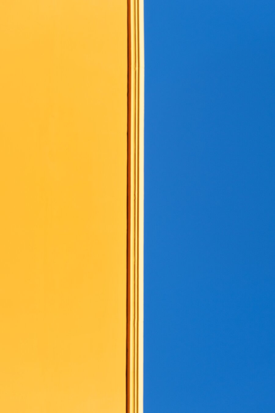This photograph is a close-up of a vertical yellow wall and blue sky. The left side is a solid, vibrant yellow/orange while the right side is a deep, solid blue. The two colours are separated by a thin, vertical roof ledge with lines running down the centre. That's it really!