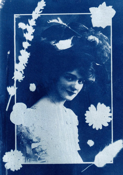 A blue-toned cyanotype portrait of a young woman in a large decorative hat, framed by white flower silhouettes. She faces slightly to the side with a gentle expression, creating a vintage, elegant composition.