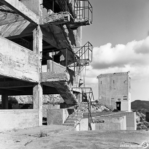 A monochrome image of an abandoned concrete structure featuring exposed staircases, crumbling walls, and a distant outbuilding. The sky is partly cloudy, enhancing the desolate atmosphere of the scene.