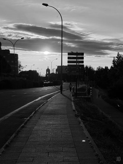 La imagen muestra una calle urbana al atardecer en blanco y negro. En primer plano se ve una acera con baldosas que avanza en perspectiva hacia el horizonte, flanqueada por farolas altas. A la derecha hay un sendero más bajo rodeado de vegetación y algunos peatones caminando. En el fondo, el sol se encuentra parcialmente cubierto por nubes y detrás se distingue la silueta de un edificio con una torre. También hay varias señales de tráfico visibles en el centro de la escena.