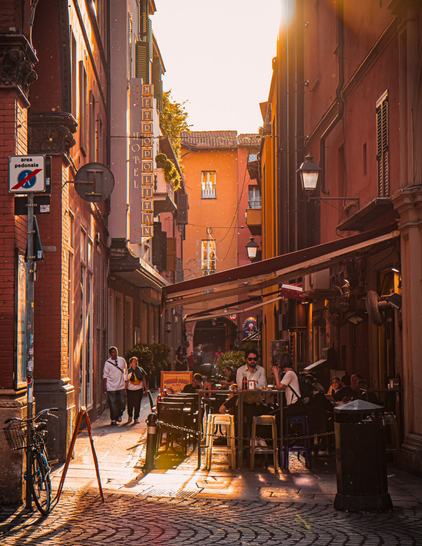 This is a photo of a narrow, sunlit Bolognese alleyway. There is warm, golden light which casts long shadows on cobblestone pavement. As is Bologna there are red and brown brick buildings with shuttered windows on both sides. Some outdoor café tables and chairs are visible with people sitting and chatting. Ther is a bicyclewhich  leans against a building on the left. 
You can see some poplar cotton in the air - it looks like glitter!