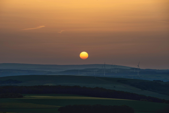 Das Bild zeigt eine weite Hügellandschaft in der Abenddämmerung unter einem warmen, atmosphärischen Himmel. Im Zentrum steht die tiefstehende, kreisrunde Sonne, die als leuchtend gelb-orangefarbene Scheibe kurz über dem Horizont schwebt und den Himmel in sanfte Nuancen von hellem Gelb bis hin zu einem gedeckten Braun-Orange taucht. Die Landschaft im Vorder- und Mittelgrund ist in dunklen, kühlen Grün- und Blautönen gehalten, wobei sich sanft geschwungene Hügelketten und bewaldete Flächen schattenhaft voneinander abheben. In der Ferne, auf einem der flachen Bergrücken, sind die filigranen Silhouetten mehrerer Windkraftanlagen zu erkennen, deren Rotorblätter sich gegen den dämmrigen Himmel abzeichnen. Ein dezenter Kondensstreifen eines Flugzeugs zieht sich als feine, helle Linie durch den oberen linken Bildbereich und verstärkt die Ruhe der Szenerie. Die gesamte Aufnahme wirkt durch den Kontrast zwischen dem warmen Himmelslicht und der dunklen Erde sehr friedlich und weitläufig.