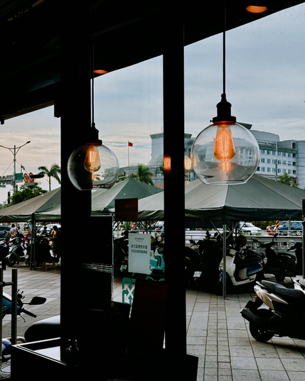 View through a window showing hanging pendant lights inside a cafe and a street scene outside with tents, parked scooters, and buildings.