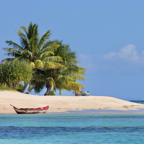 eine tropische strandlandschaft unter strahlend blauem himmel.

vorne klares, türkisblaues wasser mit sanften wellen. ein schmales, rot-weißes holzboot liegt am ufer. dahinter ein heller sandstrand, auf dem weitere kleine boote im schatten von palmen stehen. im hintergrund dichte, grüne palmen und ein wolkenloser himmel, der am rechten rand eine leichte schleierwolke zeigt.

die atmosphäre wirkt ruhig, sonnig und paradiesisch.