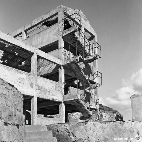 A black and white photograph of a partially constructed concrete building with exposed frameworks, irregular shapes, and a staircase. The surrounding area features rocky textures and a cloudy sky in the background, emphasizing the structure's abandoned state.