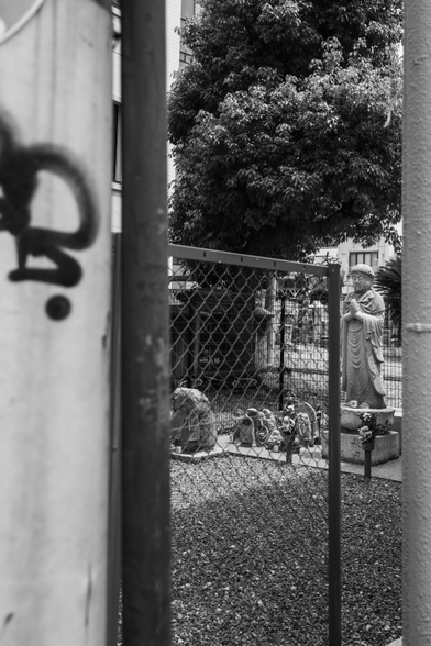 Black adn white photo of a small neighborhood Buddhist shrine in Osaka, Japan, glimpsed through a narrow gap between buildings. I believe, the tall figure on the right is a Jizō (地蔵) statue — hands pressed together in prayer, draped robes, serene expression. I took this photo at a cemetery, almost in the middle of a city. 