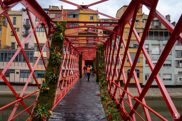 Color photo looking down the structure of an old iron pedestrian bridge with colorful low buildings on the other side. On the bridge is a person in an orange safety vest walking towards other pedestrians beyond.