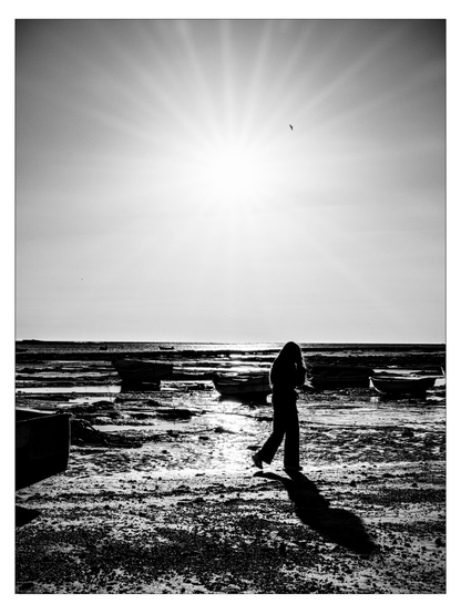 Das SW-Bild zeigt eine Person im starken Gegenlicht der Sonne am späten Nachmittag kurz vor Sonnenuntergang. Die Person spaziert am Strand, da gerade Ebbe ist, liegen einige kleine Fischerboote am trockengefallenen Strand.
