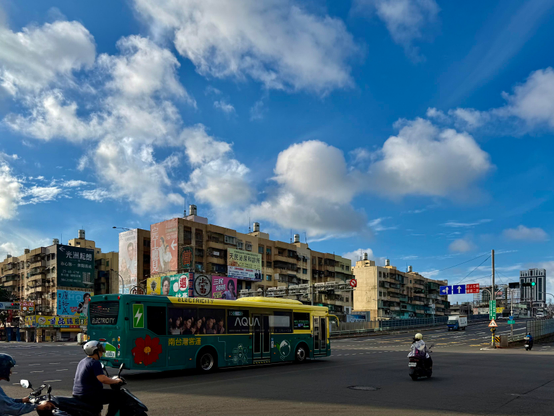 繁忙的城市街道上，公交车和摩托车在交汇处通行，背景是多层建筑和蓝天白云。