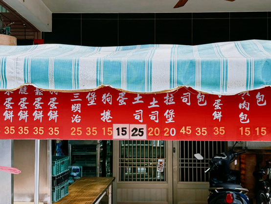 Red sign with white Chinese characters and prices displayed in yellow and black, hanging against a storefront with a blue and white striped awning.