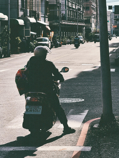 一名骑摩托车的人在街道边等待，背影遮挡，城市街景背景。