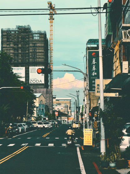 A city street scene with cars, a cyclist, and a scooter, under a blue sky with some clouds, featuring tall buildings, signage, and a construction crane.