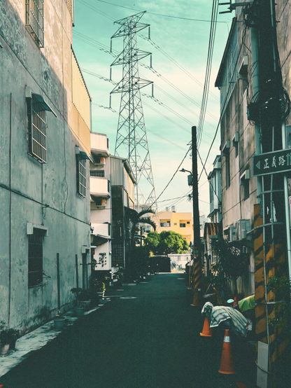 A narrow alley between tall buildings with utility poles, wires, and a large electrical tower under a partly cloudy sky.