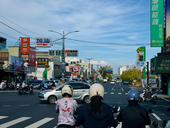People on motorcycles waiting at a busy crossroad in an urban area with various shops and traffic signs.