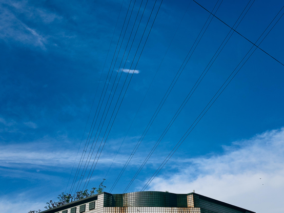 Clear blue sky with wispy clouds over a building with small plants on the roof and power lines crossing the sky.