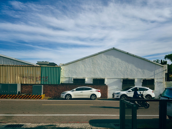 A street scene showing a white car, a motorcyclist, and a white building with three vents under a partly cloudy sky.