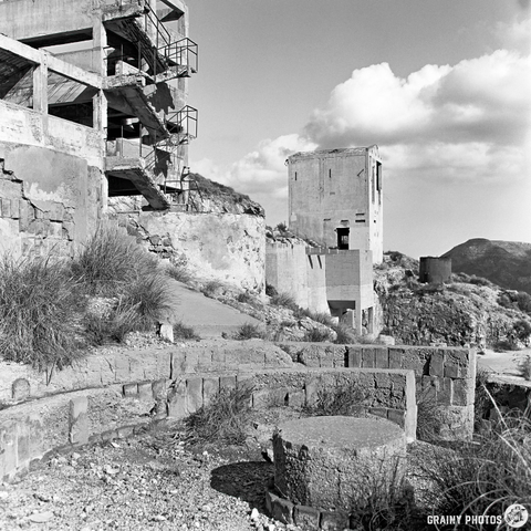 A black and white photograph showcasing a dilapidated concrete structure with a winding path, steps, and overgrown vegetation. The scene features dramatic clouds in the background and remnants of industrial architecture, evoking a sense of abandonment.