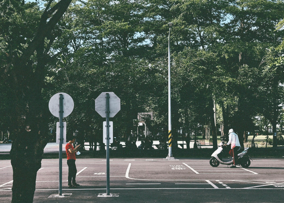 A person on a scooter rides past a parking lot with two individuals standing, one of whom is using a phone, under large trees and a street lamp.