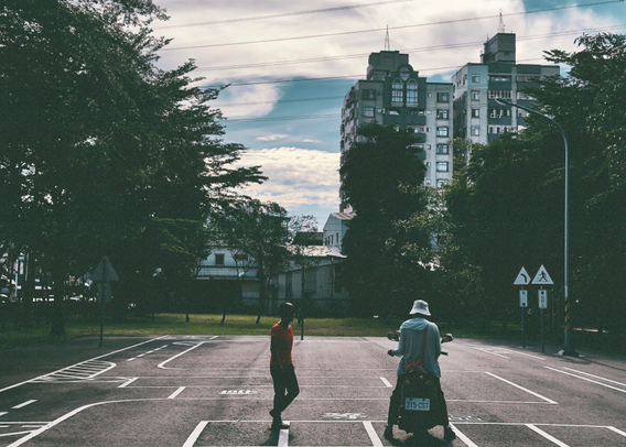 Two people, one standing and one sitting on a scooter, in an empty parking lot with trees and tall buildings in the background.