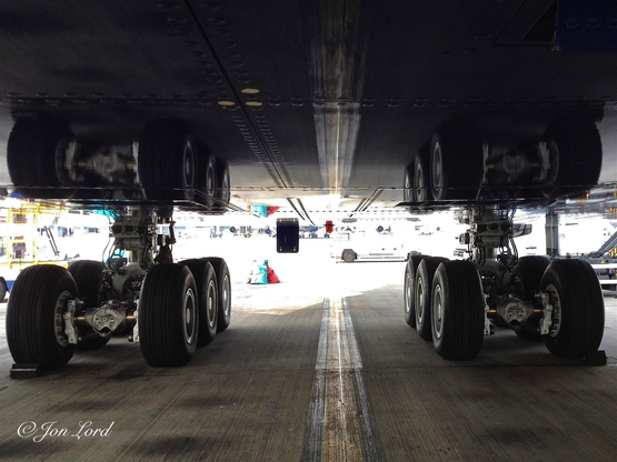 This is a colour abstract photo in landscape format of two sets of aircraft undercarriages with their image reflected in the paint work of the fuselage above. (2013)

From the base of the photo and from the centre, a thick yellow line stretches upwards and away from the camera. Either side of the line is an expanse of concrete, stained in places with a symmetrical patterns of four black patches either side of the yellow line. About two metres to the left and right of the line are two large sets of aircraft undercarriages each with six large tyres and with a single thick shaft rising up into the plane. Above is the dark blue and flat underside of the fuselage with all the tyres and undercarriage reflected in the shiny and clean surface. Beyond is bright sunlight with much of the background overexposed to white, though some of the ground equipment can be seen such as the scissors jack mechanism of a cargo loading machine.

The wheels belong to an Airbus 380 aircraft parked at Heathrow Airport in the UK. Out of view are ten further wheels. 


