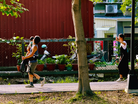 A man carries a small child while walking on a sidewalk, with a woman taking a photo or waving nearby behind a fence and trees, in a sunny outdoor setting.