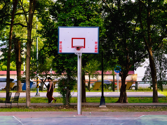 Empty basketball court with a basketball hoop, surrounded by trees and a person walking by.