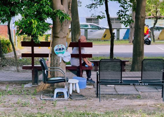 An elderly person sitting on a bench under trees in a park, with chairs and objects around, and a street visible in the background.