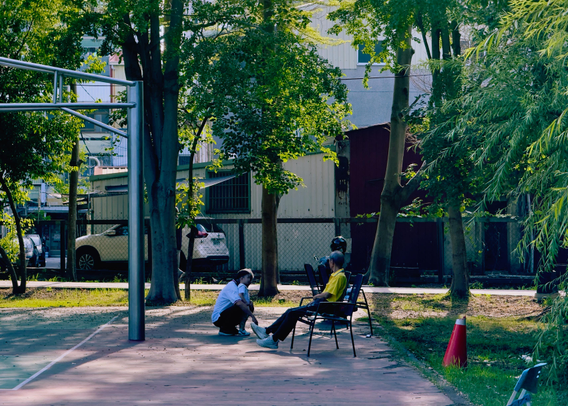 A person in a yellow shirt is sitting on a park bench while another person kneels in front of them, holding their foot, with trees and buildings in the background.
