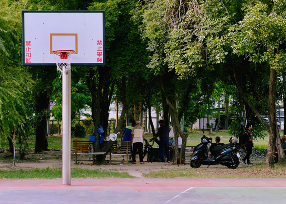 People sitting and standing under trees near a parked scooter by an outdoor basketball court. The basket's pole and backboard are visible.